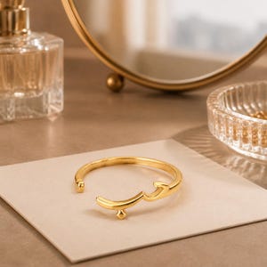May include: A gold-toned open cuff bracelet featuring a polished finish and a central Arabic script design. The metal band is circular with a small diamond-shaped accent at the base of the lettering, creating a sleek and continuous metallic form.