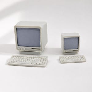 Puede incluir: Dos conjuntos de computadora en miniatura de color gris claro, cada uno compuesto por un monitor con pantalla azul claro y un teclado separado. El conjunto más grande está a la izquierda y el más pequeño a la derecha, ambos con teclas y marcos detallados.