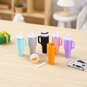 May include: A collection of miniature tumblers in light blue, silver, black, pink, orange, and purple, each featuring a side handle, a white lid, and a straw. One orange lid is detached and placed next to a single white straw on the surface.