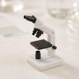 May include: A miniature white microscope featuring a black eyepiece, a rotating turret with multiple objective lenses, a square black stage with a textured surface, and a black light source component mounted on a solid white rectangular base.