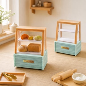 Op de afbeelding: Twee miniatuur bakkerijvitrines met lichtblauwe houten onderstellen en beige frames. Elke vitrine is voorzien van het woord PÂTISSERIE op een gouden plaatje. Eén vitrine bevat miniatuur broden en gebak op twee witte planken, de andere is leeg.