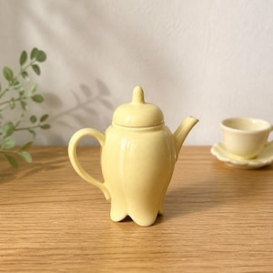 May include: A small, pale yellow ceramic teapot with a tulip-shaped body, a matching lid with a pointed finial, a curved handle, and a short spout. Beside it sits a matching small teacup on a saucer, both in the same pale yellow finish.