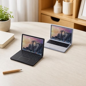 Può includere: Due repliche di computer portatili in miniatura, una in nero opaco e una in grigio chiaro, ciascuna con uno schermo che mostra un paesaggio montano, una tastiera nera e un trackpad, posizionate su una superficie chiara con una matita.