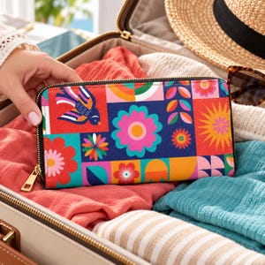 Puede incluir: Una cartera rectangular con cremallera que presenta un patrón geométrico multicolor y vibrante. El diseño incluye flores estilizadas, un pájaro y motivos de rayos de sol en tonos rosa, naranja, azul, rojo y verde sobre fondo azul oscuro.