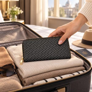 Peut inclure: Un portefeuille rectangulaire noir orné d'un motif en nid d'abeille blanc sur toute sa surface. Le portefeuille est doté d'une tirette de fermeture éclair dorée et est posé sur des vêtements pliés beiges et fauves dans une valise noire.