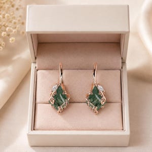 May include: A pair of rose gold drop earrings featuring a faceted green moss agate stone encased in a delicate metal leaf setting with a small clear accent stone at the top, presented inside a light beige rectangular jewelry display box.