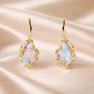 May include: A pair of gold drop earrings featuring a square-shaped iridescent opal stone set within a delicate metal frame of vine-like leaves and small clear crystals. The earrings have a curved hook closure and a polished gold finish.
