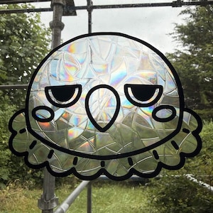 May include: A window decal featuring a stylized bird head with a rounded shape, a pointed beak, and two oval cheeks. The design is outlined in thick black lines and filled with a holographic, shattered glass pattern that reflects light.