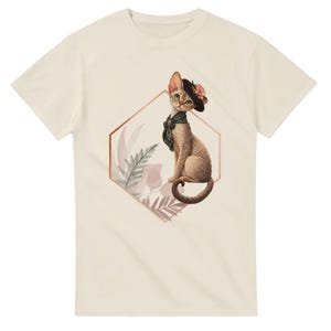 Peut inclure: Un t-shirt à manches courtes de couleur crème arborant un motif de chat portant un béret noir avec une fleur rose et une écharpe vert foncé. Le chat est placé à l'intérieur d'un cadre hexagonal cuivré avec des illustrations de fougères roses.