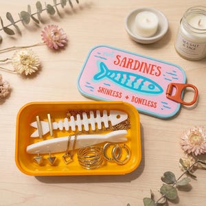 Op de afbeelding: Een rechthoekige gele opbergdoos met een lichtblauw deksel met een roze midden, een blauwgroene visafbeelding en de tekst "not SARDINES SKINLESS + BONELESS". Binnenin bevinden zich goudkleurige sieraden en witte ornamenten in de vorm van vissen.