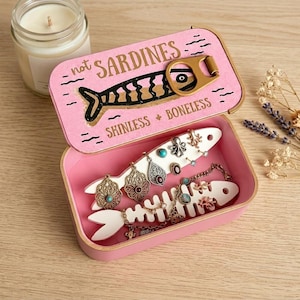 May include: A pink rectangular metal tin with a hinged lid featuring a sardine graphic and the text "not SARDINES" and "SKINLESS + BONELESS". Inside, two white fish-shaped inserts hold various metal jewelry pieces, including earrings and a chain.