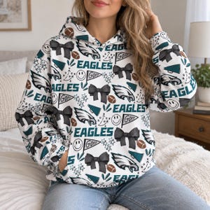 Könnte beinhalten: Ein weißer Kapuzenpullover mit einem Allover-Print aus dunkelgrünen Philadelphia Eagles-Logos, schwarzen Schleifen, braunen Footballs, türkisfarbenen Megafonen, Smileys und Wimpeln mit dem Text GO. Der Hoodie verfügt über eine Kängurutasche.