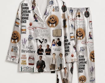 Peut inclure: Un ensemble de pyjama en satin blanc composé d'une chemise à manches longues boutonnée et d'un pantalon assorti. Le tissu est orné d'un collage de graphismes The Vampire Diaries, incluant des illustrations de personnages et du texte.