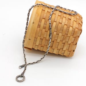 May include: A silver chain necklace featuring a textured, interlocking link design with a circular pendant at the end. The chain has a darkened, oxidized finish in the crevices of the links, providing a high-contrast, metallic appearance.