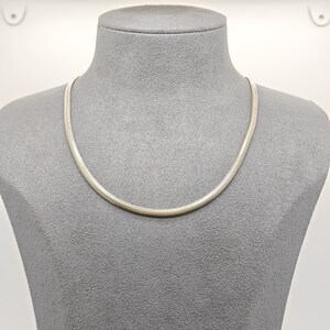 May include: A silver-toned snake chain necklace featuring a smooth, flexible, and tightly woven metallic texture. The chain has a rounded profile and a polished finish, resting in a simple circular shape around the neck of the display form.
