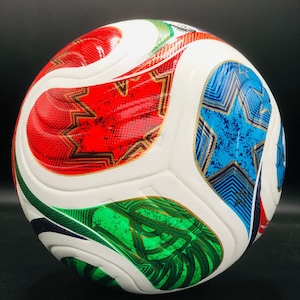 May include: A white soccer ball featuring a multi-colored graphic design with red, blue, and green star-shaped panels. The surface has a textured finish with gold-colored outlines surrounding the vibrant, patterned shapes across the sphere.