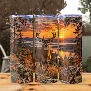 May include: A cylindrical tumbler featuring a wrap-around graphic of a buck with large antlers standing in a forest at sunrise. The scene includes orange and yellow clouds, a calm lake, pine trees, and rocky terrain with autumn-coloured foliage.