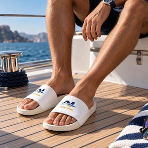 Könnte beinhalten: Ein Paar weiße Schlappen mit einem blau-orangen Grafiklogo auf dem Riemen und dem darunter in Gelb gedruckten Text BOAT NAME. Die Sandalen verfügen über ein geformtes Fußbett und einen breiten Riemen über dem Fußrücken.