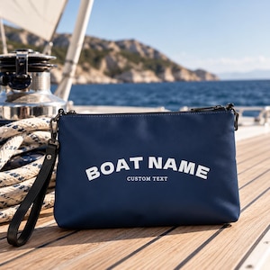 Könnte beinhalten: Eine marineblaue, rechteckige Reißverschlusstasche mit schwarzer Handschlaufe. Auf der Vorderseite befindet sich der weiße Schriftzug "BOAT NAME" in fetter, gebogener Schrift über dem kleineren weißen Text "CUSTOM TEXT" auf der matten Oberfläche.