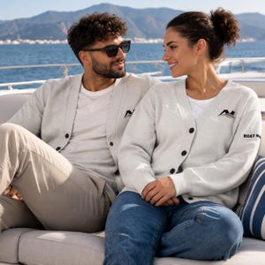 May include: Two light gray V-neck button-down cardigans featuring a black and blue logo on the left chest. The cardigan on the right includes the text BOAT NA on the sleeve. Both garments are worn over white t-shirts with casual trousers.