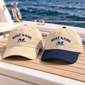 Op de afbeelding: Twee honkbalpetten in beige, één met een bijpassende beige klep en één met een marineblauwe klep. Beide petten zijn voorzien van de tekst BOAT NAME in marineblauwe letters boven een gestileerd marineblauw en goud grafisch logo.
