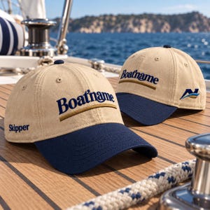Op de afbeelding: Twee beige honkbalpetten met marineblauwe kleppen. Op de voorkant van de petten staat de tekst "Boatname" in marineblauw geborduurd boven een gouden horizontale lijn. Het woord "Skipper" is in marineblauw op de zijkant geborduurd.