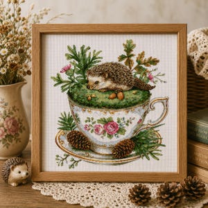 Könnte beinhalten: Ein gerahmtes Kreuzstich-Stickbild mit einem braunen Igel, der in einer weißen, mit rosa Rosen verzierten Teetasse ruht. Die Tasse steht auf einer passenden Untertasse, umgeben von grünem Laub, Tannennadeln und braunen Tannenzapfen auf weißem Stoff.