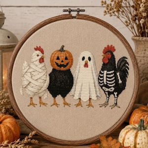 May include: A circular wooden embroidery hoop containing a cross-stitch design of four chickens in Halloween costumes. The chickens are dressed as a mummy, a pumpkin, a ghost, and a skeleton, all stitched onto a light beige fabric background.