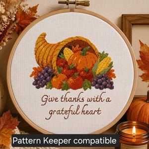 Könnte beinhalten: Ein runder Stickrahmen aus Holz mit einem Kreuzstichmotiv, das ein Füllhorn gefüllt mit Kürbissen, Mais, Weintrauben und Herbstblättern zeigt. Unter dem Erntemotiv steht der Text, Give thanks with a grateful heart.