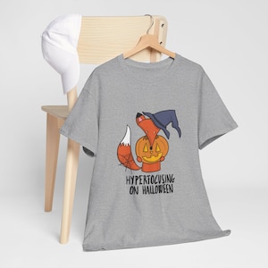 May include: A heather gray t-shirt featuring a graphic of an orange fox wearing a dark blue witch hat and holding a carved jack-o'-lantern pumpkin. The fox tail has a spiderweb design. Below the graphic is the text "HYPERFOCUSING ON HALLOWEEN".