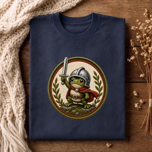 Könnte beinhalten: Ein marineblaues T-Shirt mit einem kreisförmigen Motiv eines grünen Frosches, der einen silbernen Helm und einen roten Umhang trägt und ein Schwert hält. Der Frosch steht auf einem felsigen Untergrund, umrahmt von grünen Lorbeerblättern auf cremefarbenem Grund.