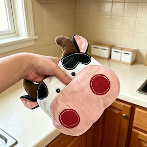May include: A cow-shaped fabric pot holder featuring a white face with black patches, brown plush horns, pink inner ears, and a large pink snout with two dark red circular nostrils. The item has small black button eyes and stitched detailing.