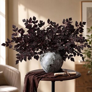 May include: A round, textured gray and white stone-like vase holds a dense arrangement of dark purple, serrated-edged leaves. The vase sits on a small, dark brown circular table draped with a piece of dark brown fabric and a rolled parchment scroll.