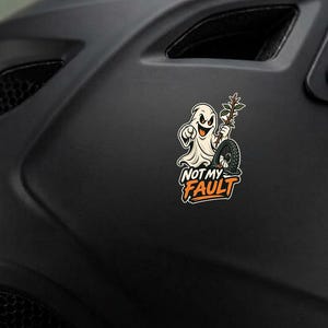 Puede incluir: Un casco de motocicleta negro mate con una pegatina que muestra un fantasma de dibujos animados señalando hacia adelante mientras sostiene una rama junto a un neumático. La pegatina incluye el texto NOT MY FAULT en letras naranjas sobre fondo blanco.