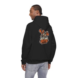 Puede incluir: Una sudadera negra con capucha que presenta un gráfico en la espalda de un murciélago de dibujos animados con alas naranjas y garras afiladas. Debajo del murciélago, el texto "JUST ONE BITE" en naranja, con "COME BACK FOR" escrito debajo.