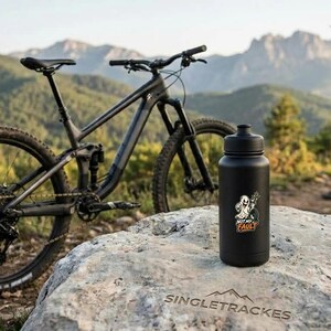 Puede incluir: Una botella de agua negra mate con tapa de rosca y una pegatina gráfica que muestra un personaje fantasma y el texto "NOT MY FAULT" colocada sobre una roca grande, con una bicicleta de montaña y una persona al fondo.