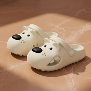 May include: A pair of off-white slip-on clogs featuring a cartoon dog face design. The shoes have a rounded toe, a black nose, small black eyes, and side cutouts that reveal white teeth. A pivoting heel strap is attached to each side of the shoe.