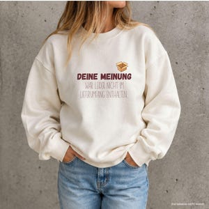 Könnte beinhalten: Ein cremefarbenes Sweatshirt mit Rundhalsausschnitt, das eine Grafik eines kleinen braunen Kartons und den Text "DEINE MEINUNG WAR LEIDER NICHT IM LIEFERUMFANG ENTHALTEN" zeigt. Das Sweatshirt hat lange Ärmel mit gerippten Bündchen und einen Rundhalsausschnitt.
