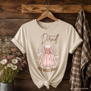 Könnte beinhalten: Ein beiges Kurzarm-T-Shirt mit einer Grafik eines rosa Dirndls, umgeben von bunten Wildblumen. Der Text "Ersatz Dirndl" ist über dem Kleid aufgedruckt, und der Text "DER WILLE ZÄHLT" befindet sich unterhalb der Grafik des Kleides.