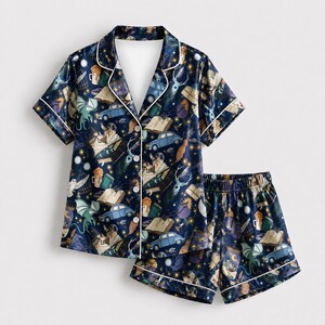 May include: A dark blue pajama set featuring a short-sleeved button-down shirt and matching shorts. The fabric displays a repeating pattern of light blue cars, open books, green dragons, white owls, and golden stars against a navy background.