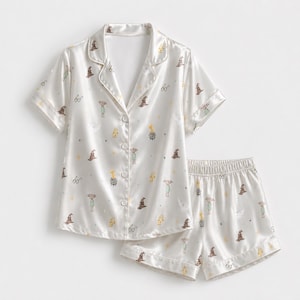 May include: A cream-colored satin pajama set consisting of a short-sleeved button-down shirt with a notched collar and matching elastic-waist shorts. The fabric features a repeating pattern of small brown hats, glasses, cauldrons, and figures.