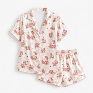 Op de afbeelding: Een tweedelige pyjamaset in een lichte crèmekleur met een herhalend patroon van Hello Kitty-figuren, roze en oranje bloemtrossen en kleine vierpuntige sterren. De top heeft een reverskraag en korte mouwen met knoopjes aan de voorzijde.