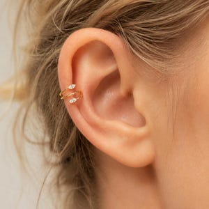 May include: A gold ear cuff featuring a delicate spiral design that wraps around the cartilage. The ends of the spiral are accented with two small, clear marquise-cut stones, providing a subtle sparkle to this minimalist piece of jewelry.