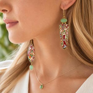Puede incluir: Pendientes colgantes con monturas de metal dorado y patrones de remolinos multicolores en rojo, verde y blanco, acentuados por pequeñas piedras ovaladas de color verde claro. Un collar de cadena dorada a juego con un colgante de piedra verde.