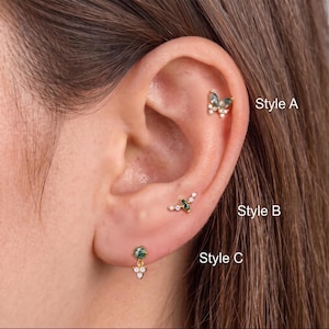 May include: Three gold-tone earrings with green moss agate stones and clear crystals. Style A is a butterfly shape, Style B is a marquise-shaped stone with small crystals, and Style C is a round stone with a dangling crystal cluster. Text labels included.