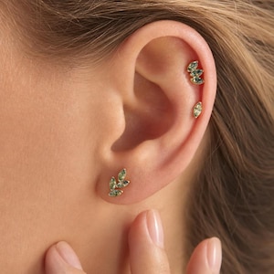 May include: A set of gold-toned earrings featuring marquise-cut green gemstones. One earring is a leaf-shaped cluster of three stones worn on the earlobe, while the upper ear cartilage features two separate piercings with individual green stones.