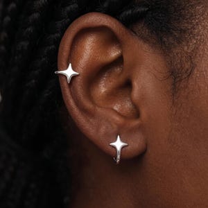 Può includere: Due orecchini color argento a forma di stella a quattro punte. Un orecchino è posizionato sul lobo, mentre l'altro si trova più in alto sulla cartilagine. Entrambi i pezzi presentano una finitura metallica liscia e lucida con punte simmetriche.