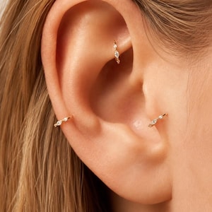 May include: Three gold hoop earrings featuring small, clear, marquise-cut and round-cut gemstones. The hoops are positioned in the rook, tragus, and helix areas of the ear, showcasing a polished metal finish and delicate, sparkling stone settings.