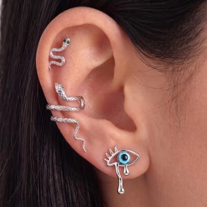 May include: A set of silver-toned earrings featuring two snake-shaped pieces with small green accents and one eye-shaped stud earring with a blue center and a teardrop detail, all worn on the cartilage and lobe of a single ear.