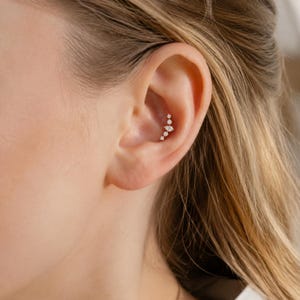 Könnte beinhalten: Ein goldfarbener Knorpel-Ohrstecker mit einer vertikalen Anordnung aus vier kleinen, klaren, rund geschliffenen Steinen und einem zentralen marquise-geschliffenen Stein, der in der Ohrmuschel sitzt und ein zartes, lineares Design bildet.